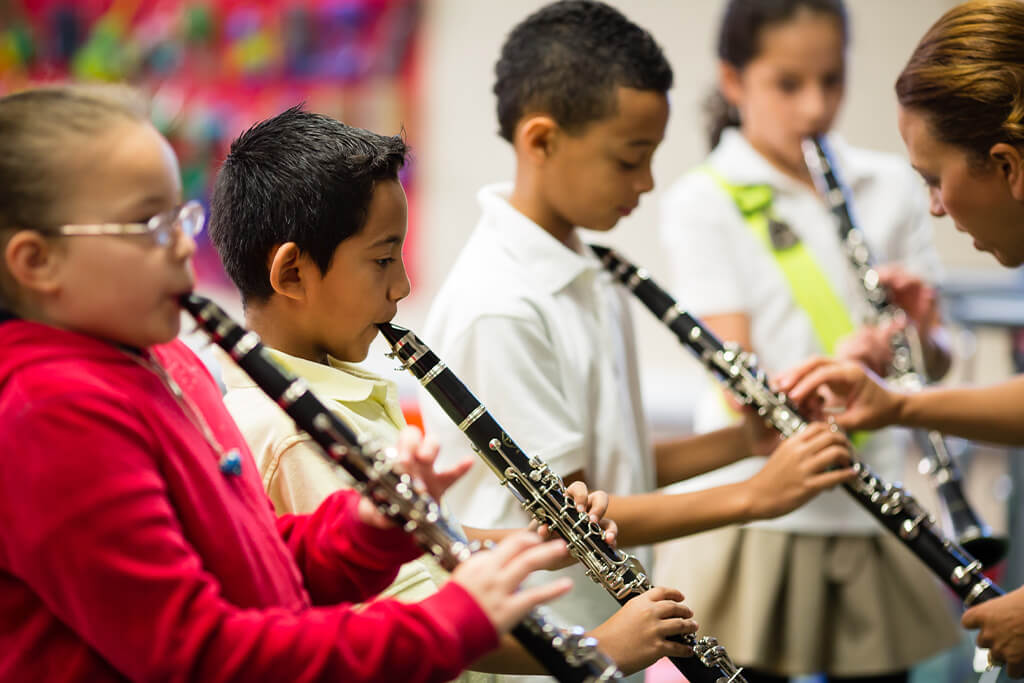 Row of students playing clarinets in a classroom, with a teacher guiding a student's hand placement.