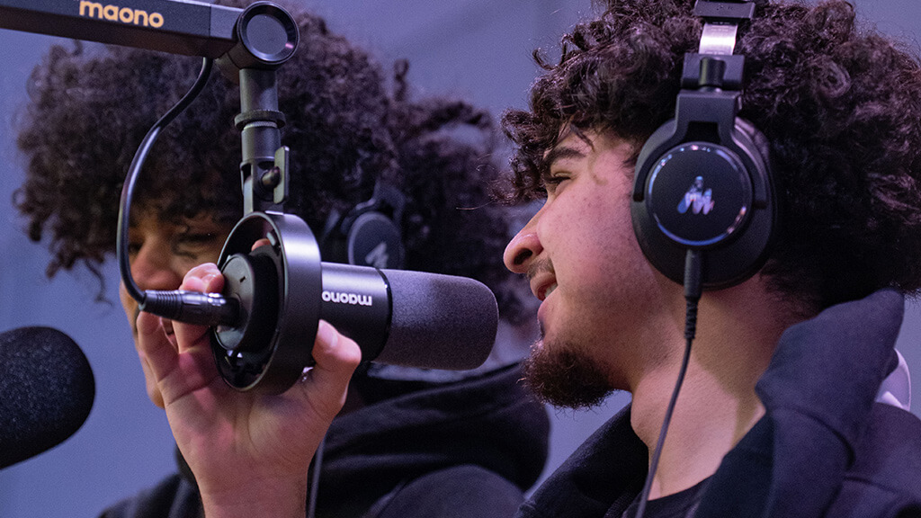 Young musician in headphones speaks into a studio microphone at a DJ Project, another participant at a mic in the background.