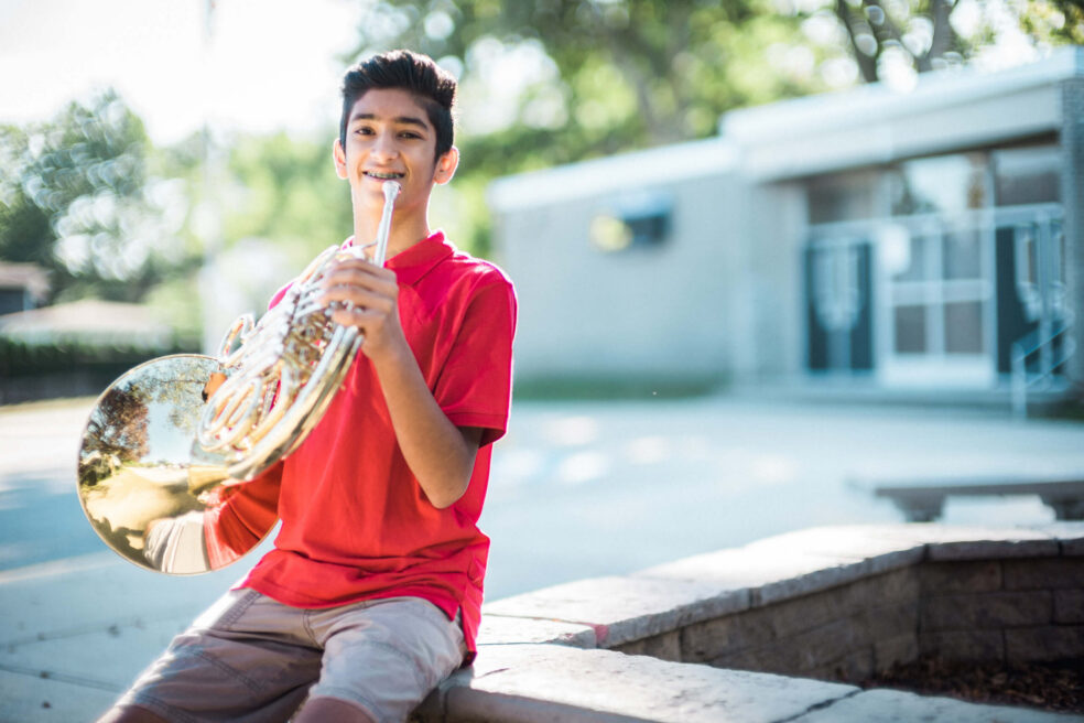 Middle school student holds a French horn outside his school.