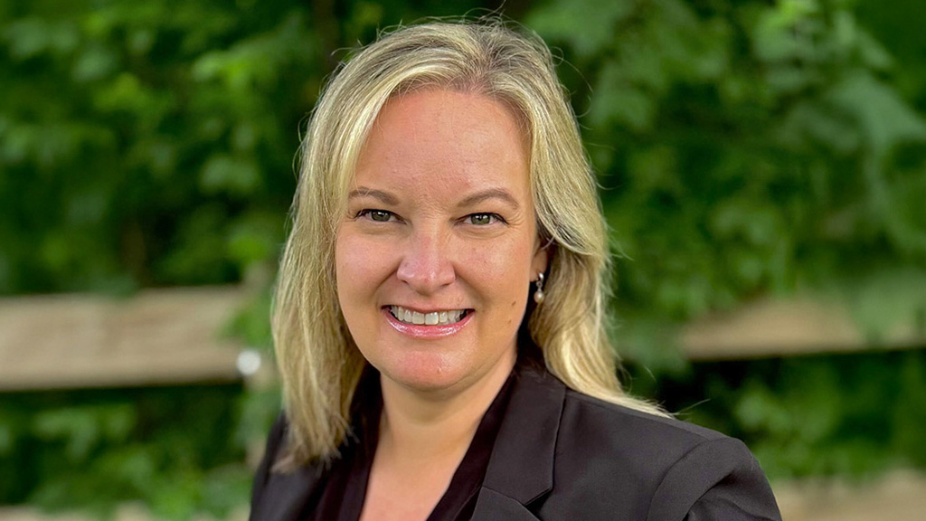 Professional headshot of a Katherine Palmer in a dark blazer, outdoors with a green foliage background.