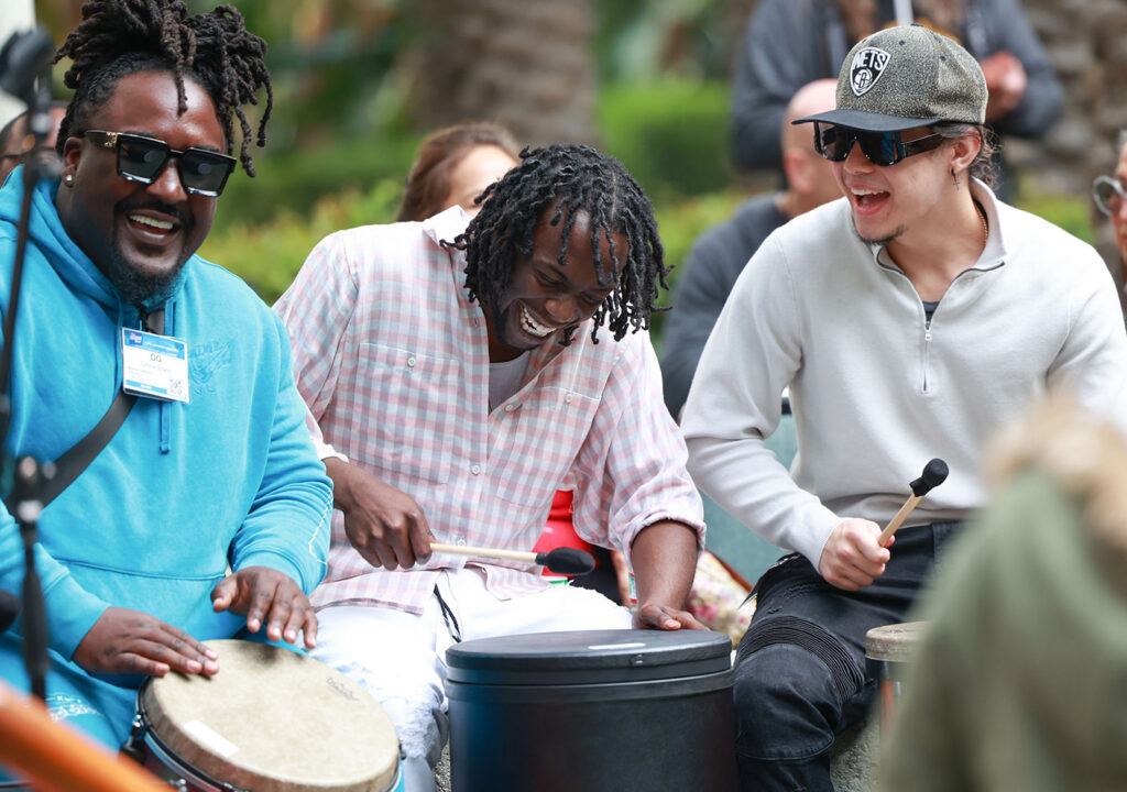 A group of men laugh and smile while playing percussion instruments with mallets, seated together outdoors with an audience visible in the background