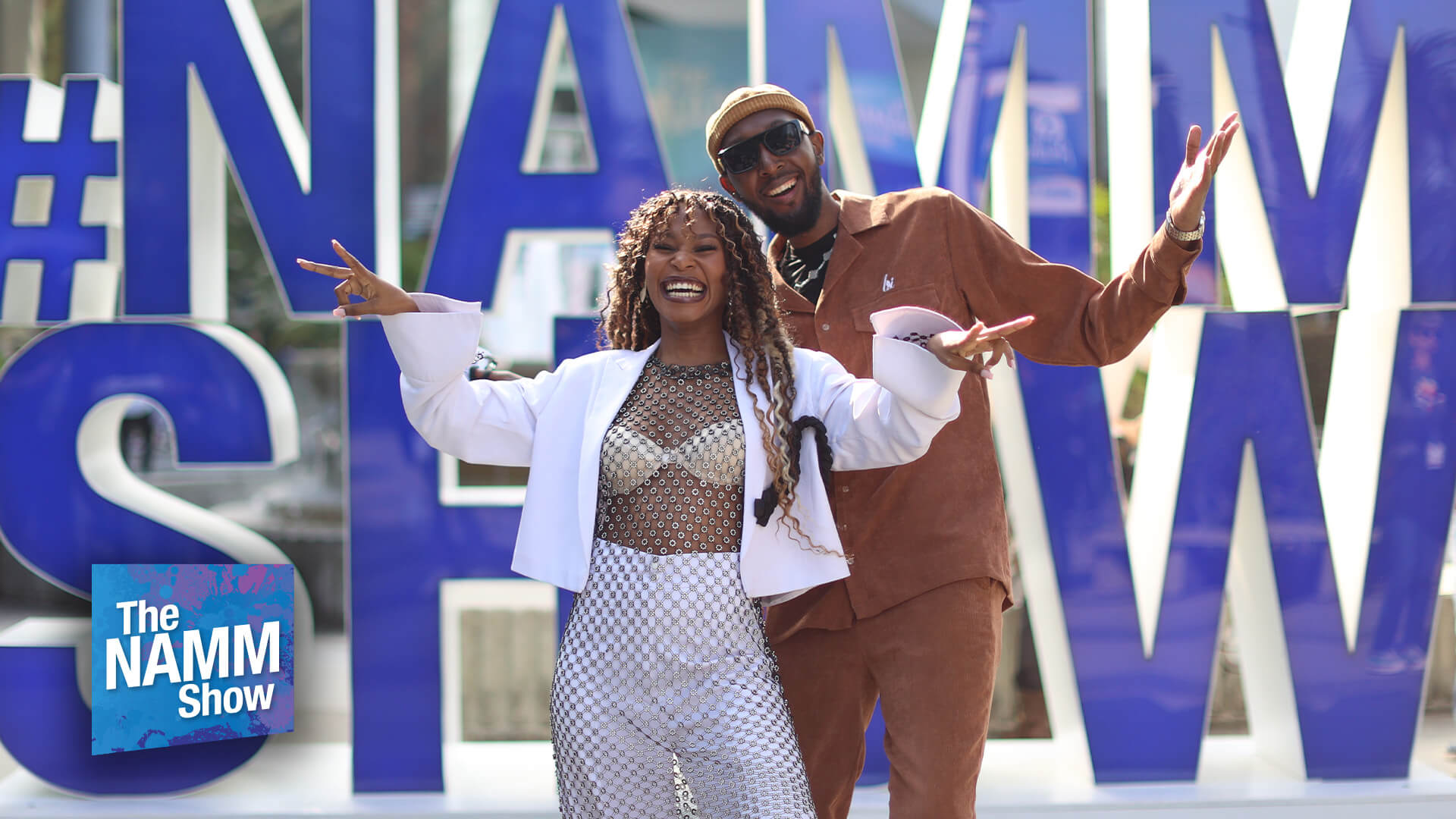 A man and woman pose energetically, smiling and gesturing with their hands, in front of a large #NAMMSHOW letter sign. The 'The NAMM Show' logo appears in the bottom left corner