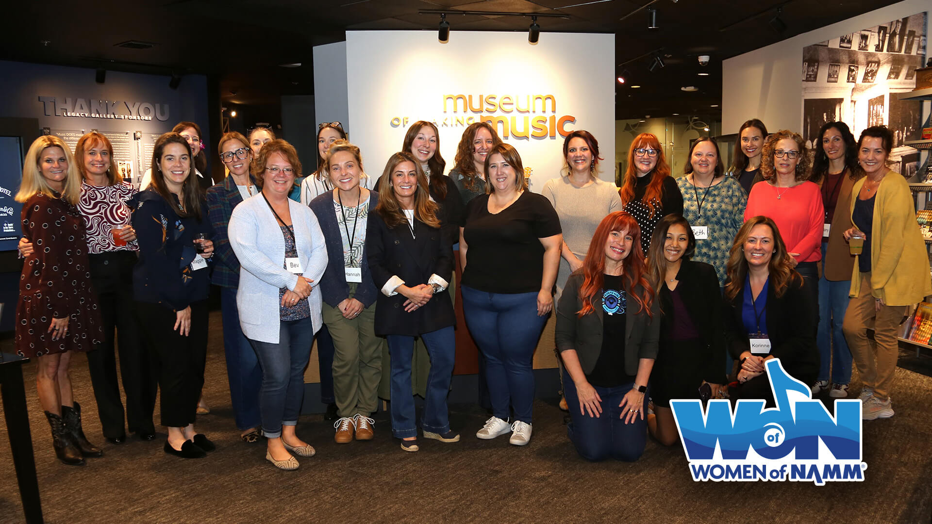 A large group of women pose together for a photo inside the Museum of Making Music, with the museum's illuminated sign visible behind them. Some wear name tags. The 'Women of NAMM' logo appears in the bottom right corner.