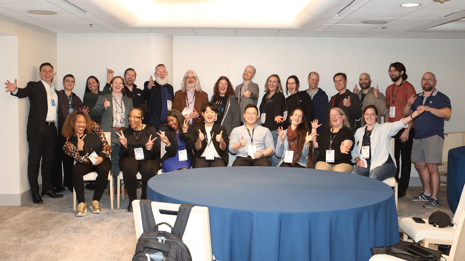 A large group of NAMM Show conference attendees pose energetically around a round table with a blue tablecloth, smiling and gesturing with peace signs and thumbs up