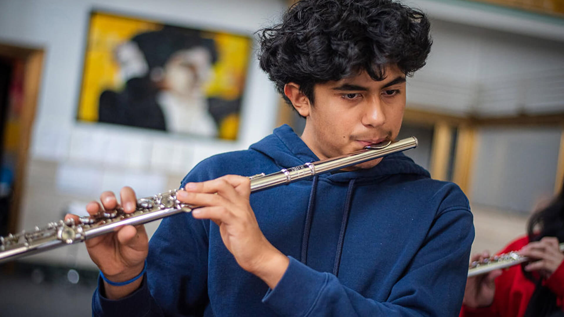 A teenage student wearing a navy blue hoodie plays flute with focused concentration, with another musician partially visible in the background