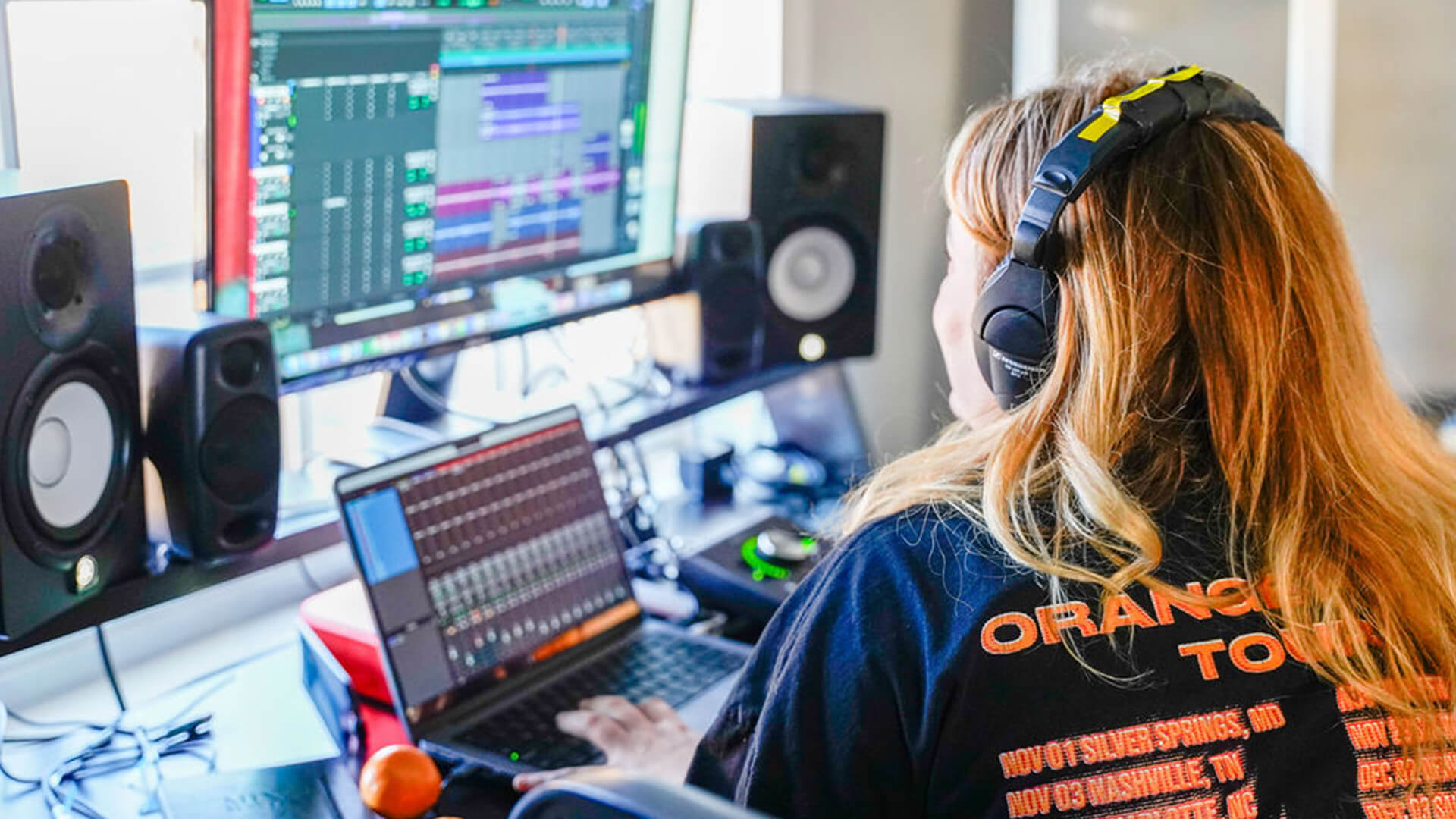 A woman wearing headphones and a tour t-shirt works at a music production desk with a laptop and monitor displaying audio editing software, surrounded by studio speakers.