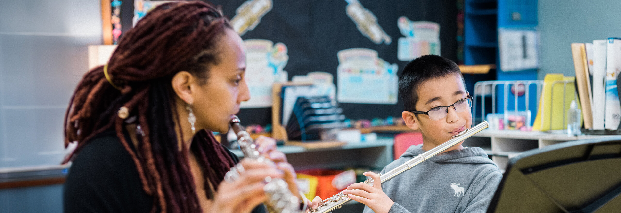 A music teacher plays flute alongside a young student who is also playing flute, both reading from a music stand in a classroom decorated with instrument posters
