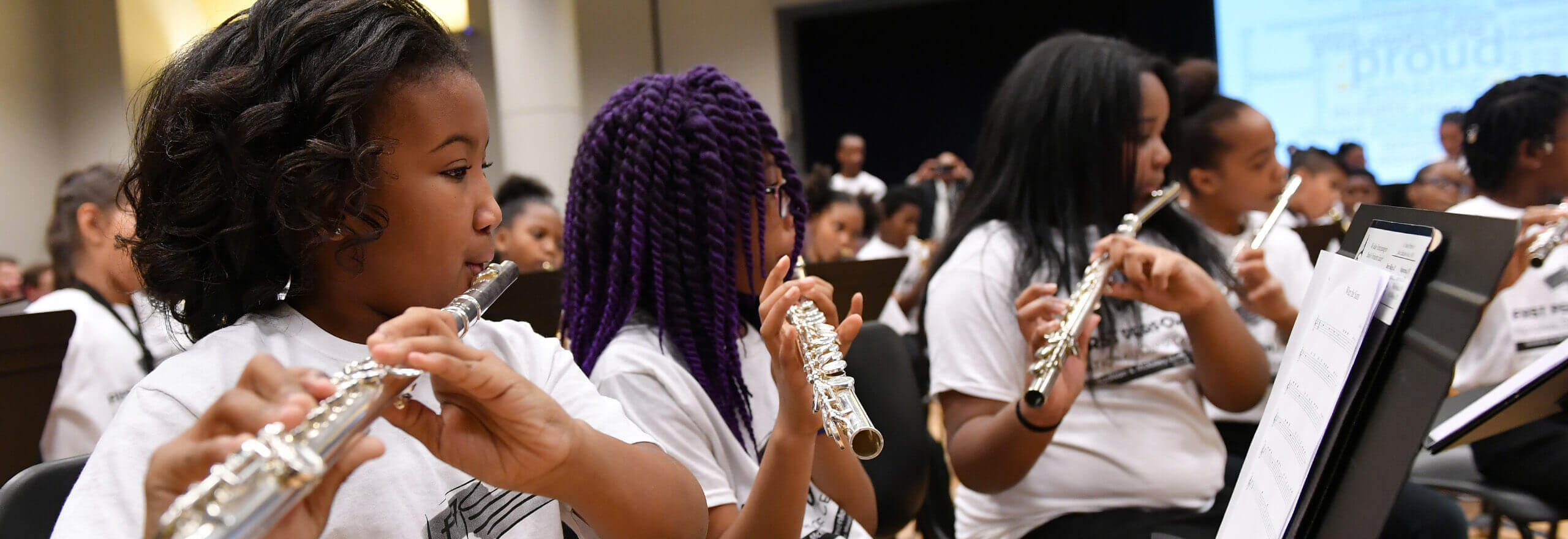 Close-up of young students playing flutes during a band performance, all wearing matching white t-shirts and reading sheet music from stands. Additional student musicians are visible in the background