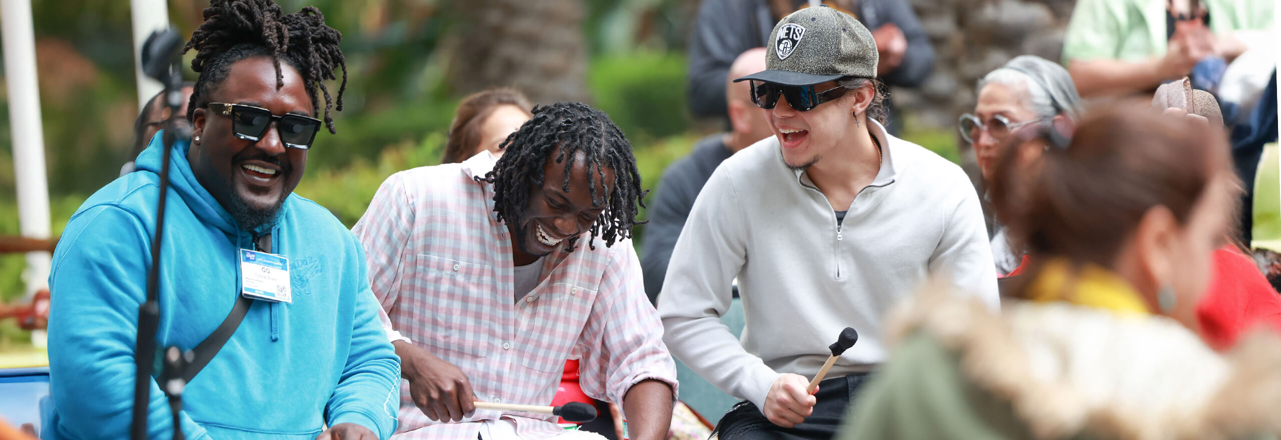 A group of men laugh and smile while playing percussion instruments with mallets, seated together outdoors with an audience visible in the background