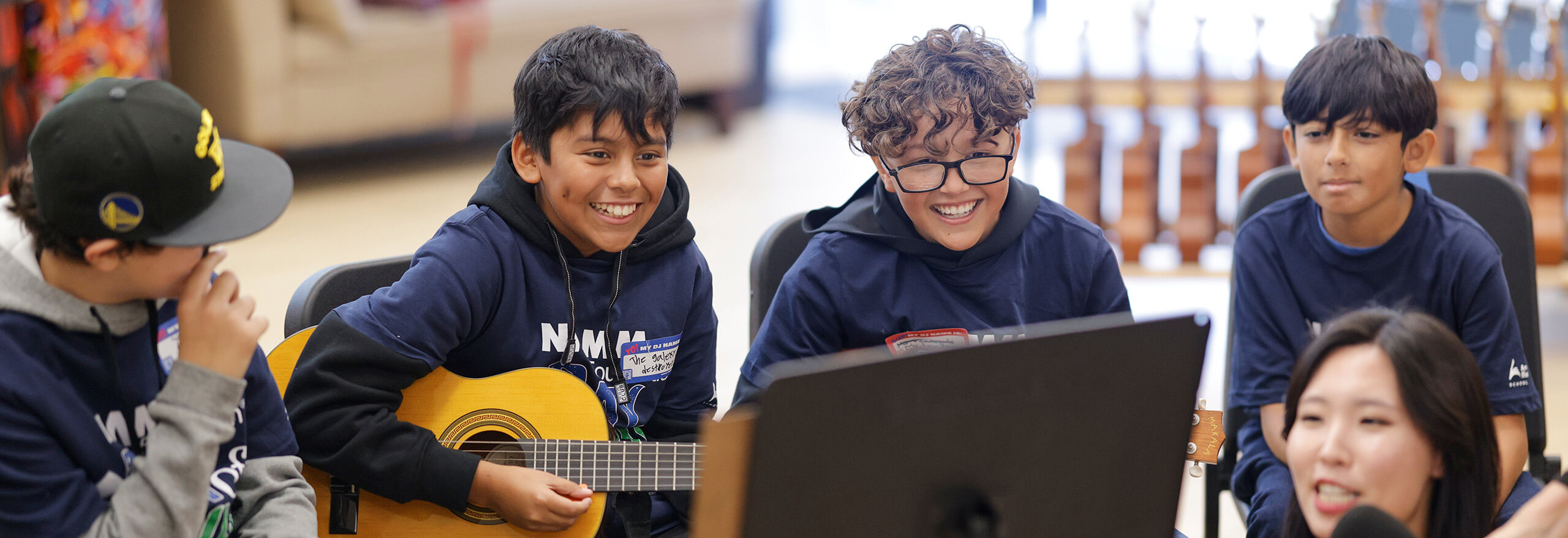 A group of smiling students wearing navy NAMM hoodies gather around a sheet music stand, with one boy playing an acoustic guitar. Rows of guitars are visible on display in the background