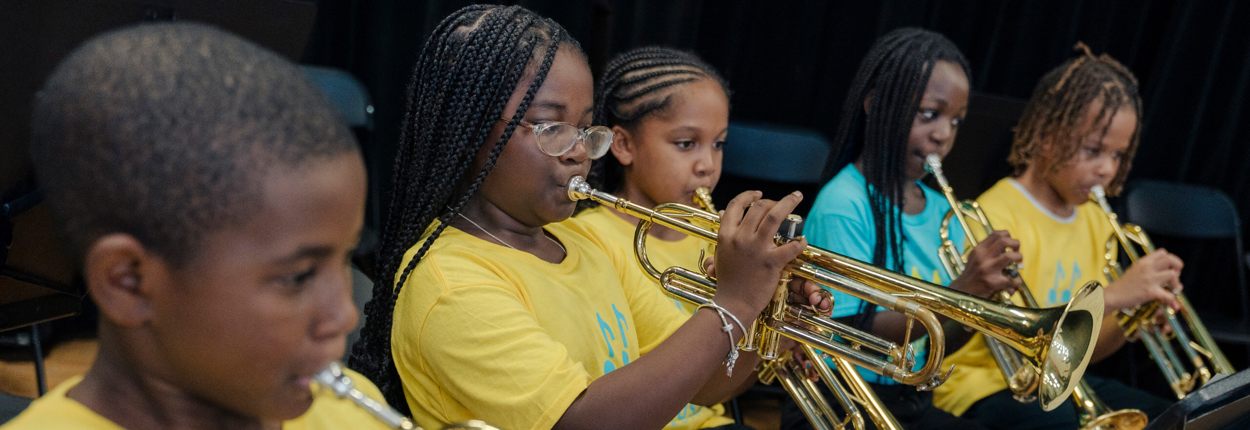 A row of young students, mostly wearing matching yellow t-shirts, play trumpets together during a band performance or rehearsal