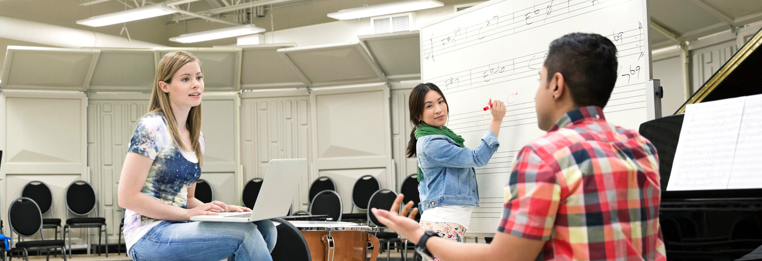 A woman writes music notation on a whiteboard while two other students, one holding a laptop and one gesturing while talking, discuss the composition in a music rehearsal room with a piano nearby