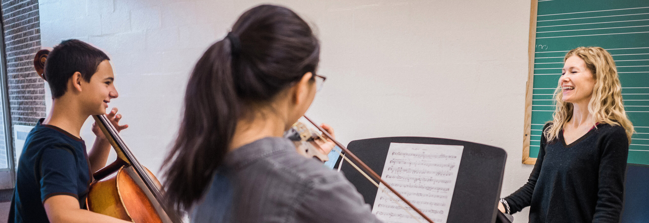 A boy plays cello and a girl plays violin while reading sheet music, smiling and interacting with their teacher, who stands nearby in front of a chalkboard in a music classroom