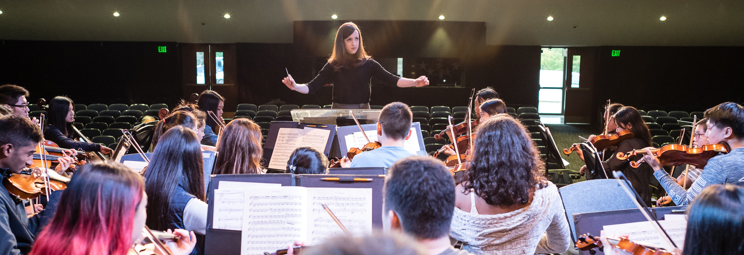 A conductor stands with arms raised, leading a student string orchestra during a rehearsal in an auditorium, with rows of empty seats visible in the background