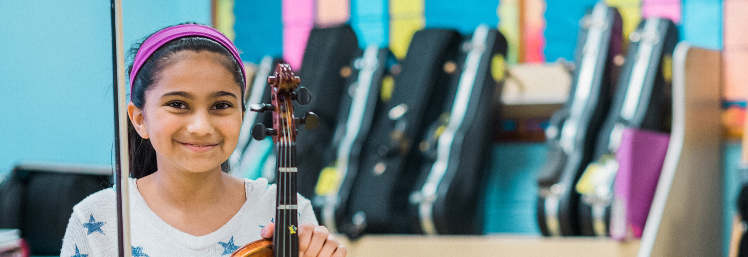 A smiling student sits in a music classroom holding a violin and bow, with instrument cases and colorful classroom displays in the background.