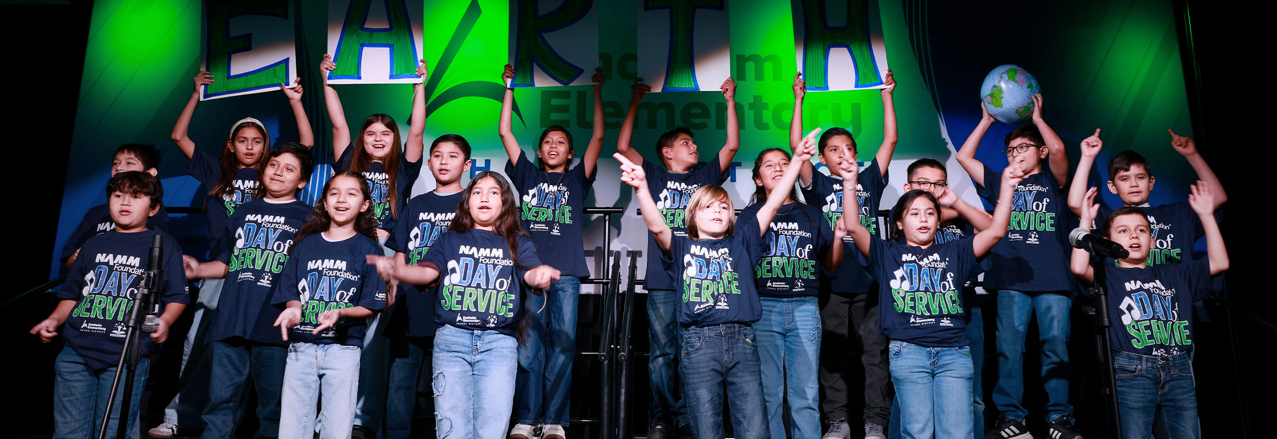 Elementary students perform together onstage during a NAMM Foundation Day of Service event, singing and raising their hands in matching shirts.