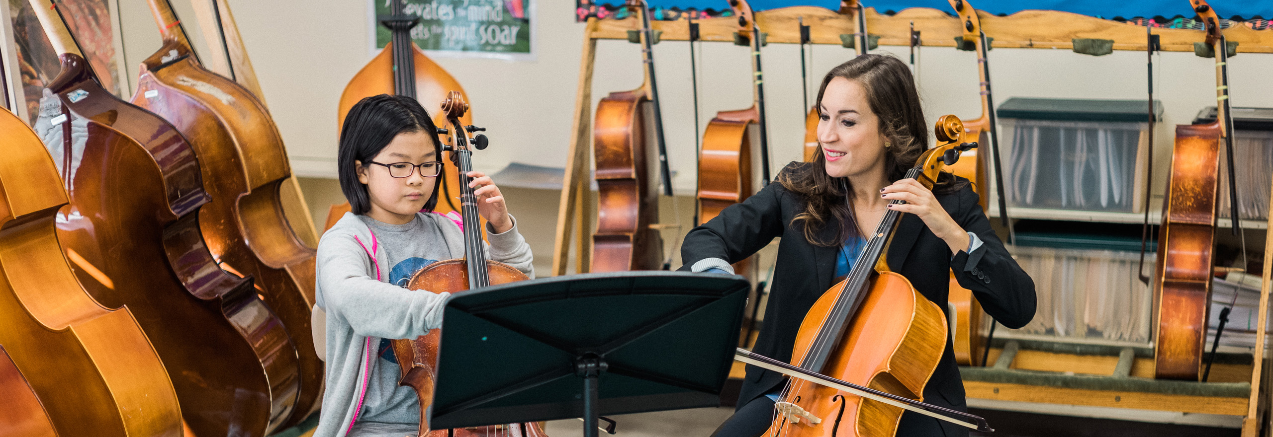 A young student plays cello during a music lesson, guided by an instructor, with several cellos visible in the classroom background.